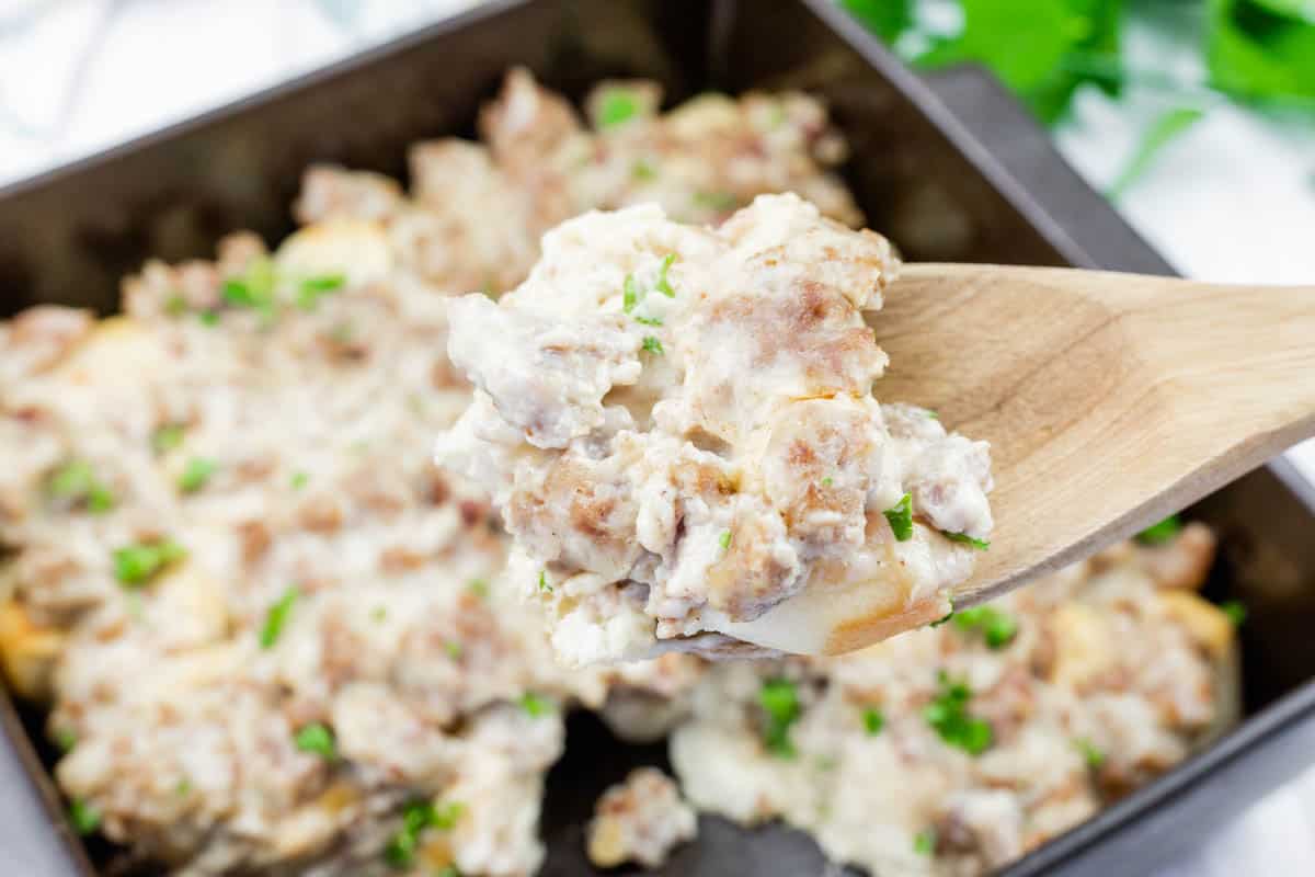 A wooden spoon holds a portion of biscuits and sausage gravy, topped with chopped parsley, over a baking dish filled with the same dish.