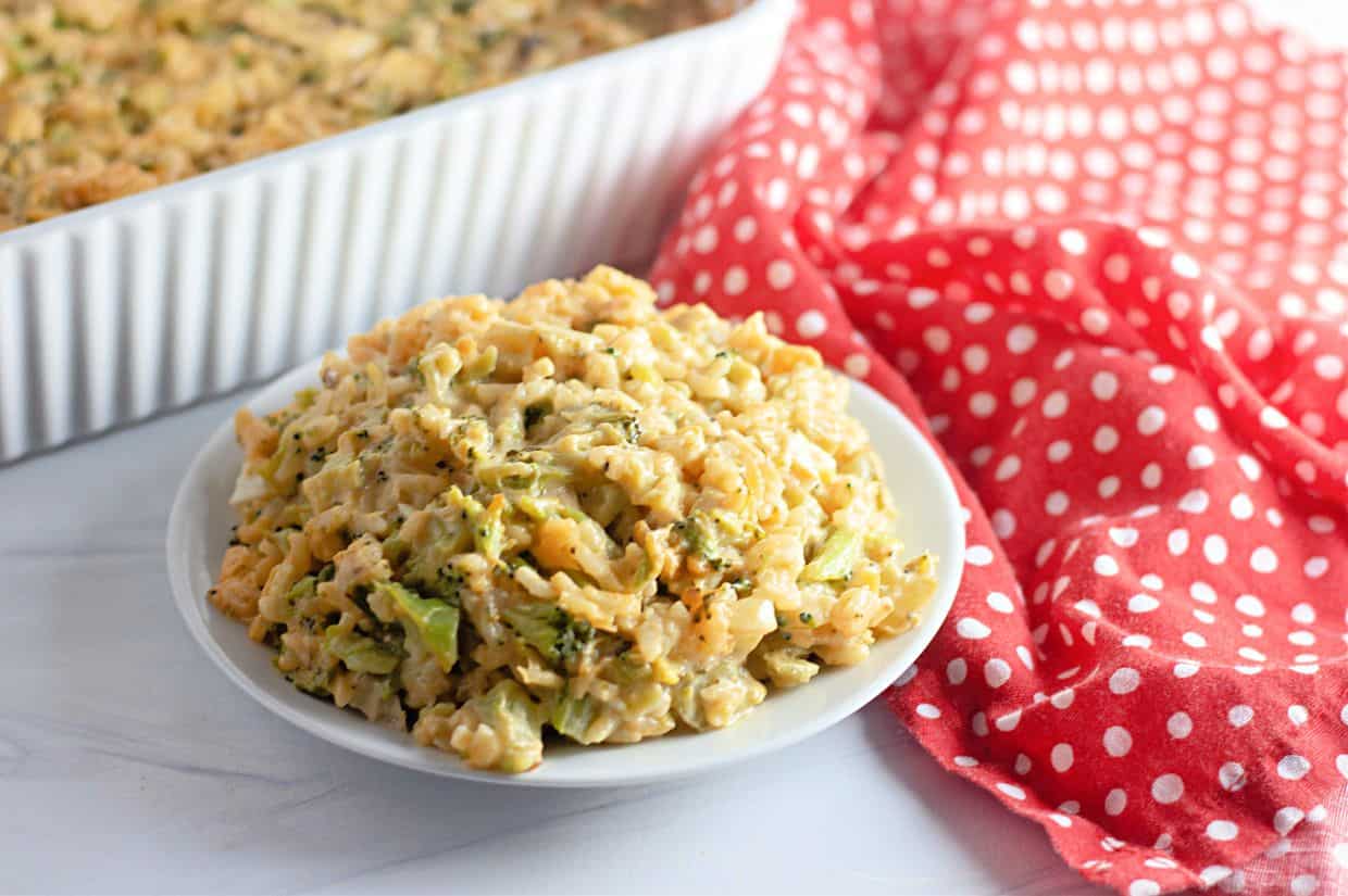 A plate of Easy Cheesy Broccoli Rice Casserole sits on a white surface next to a red and white polka dot cloth, with a baking dish in the background.