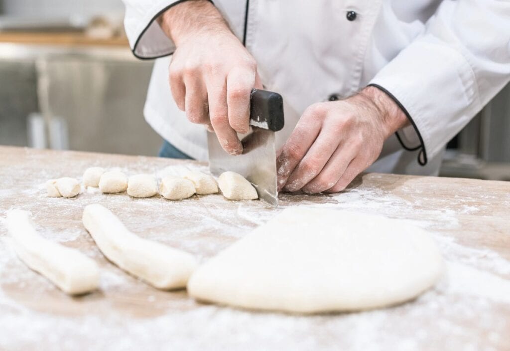 A person in a chef's coat uses a bench scraper to cut small pieces from rolled dough, with flour and dough scattered on a wooden surface.