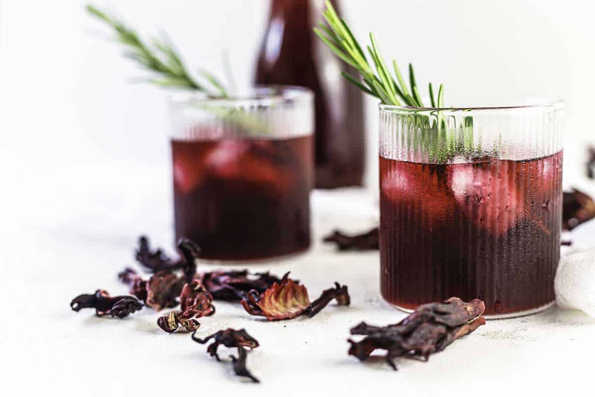 Two glasses of iced hibiscus tea garnished with rosemary sprigs, with dried hibiscus flowers scattered on a white surface. A bottle of the drink is in the background.