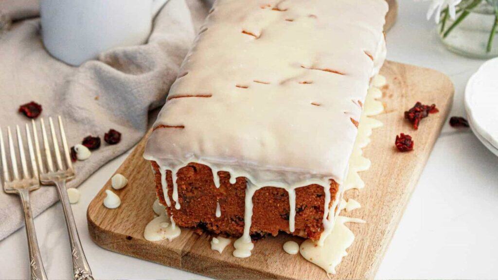 A rectangular loaf cake with white icing drips sits on a wooden board, surrounded by forks and dried cranberries.