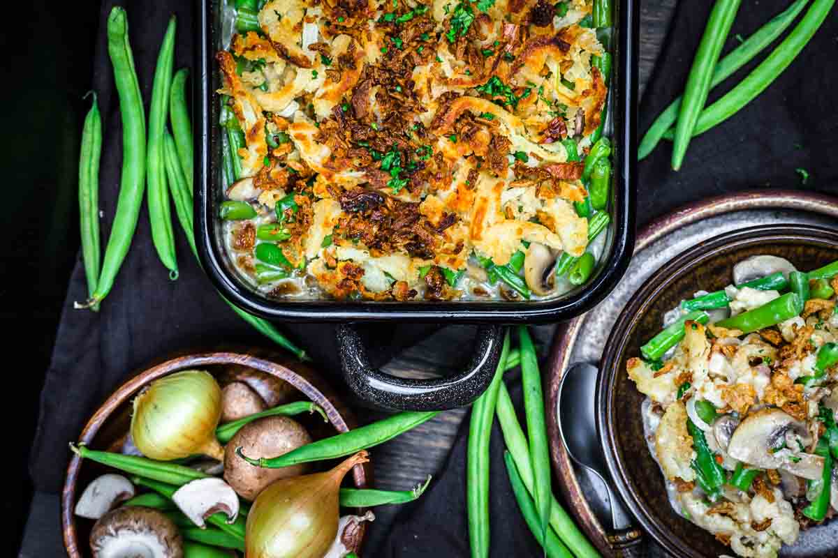 A baked green bean casserole topped with crispy onions sits next to fresh green beans, onions, mushrooms, and a plate serving of the casserole.