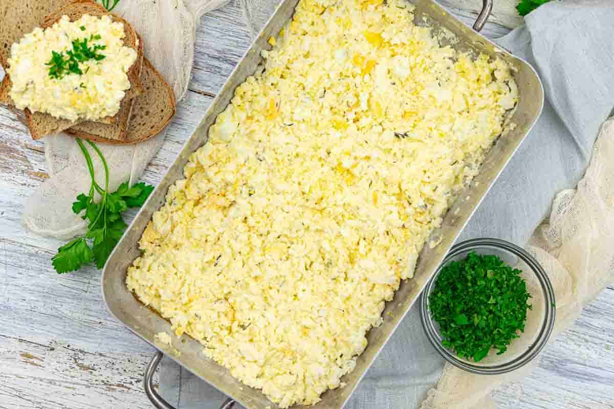 Rectangular metal dish filled with egg casserole mixture, next to a slice of bread with egg mixture, a bowl of chopped parsley, and fresh parsley on a wooden surface.