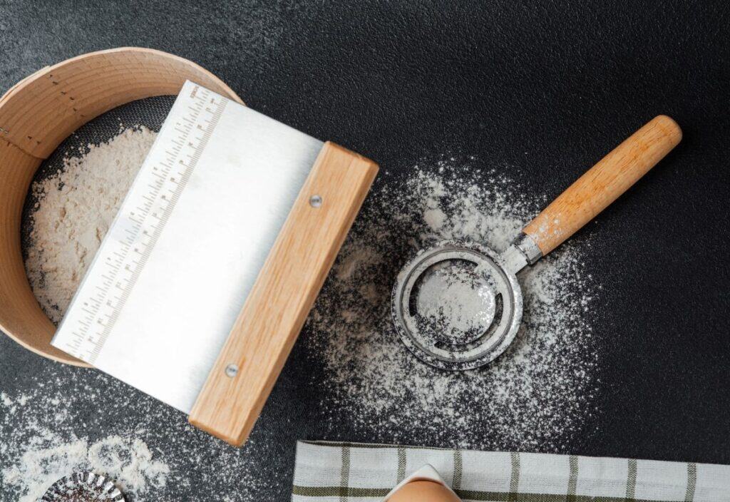 A flour sifter and a metal bench scraper with wooden handles rest on a dark, flour-dusted surface, beside a striped cloth and a single egg.