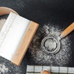 A flour sifter and a metal bench scraper with wooden handles rest on a dark, flour-dusted surface, beside a striped cloth and a single egg.