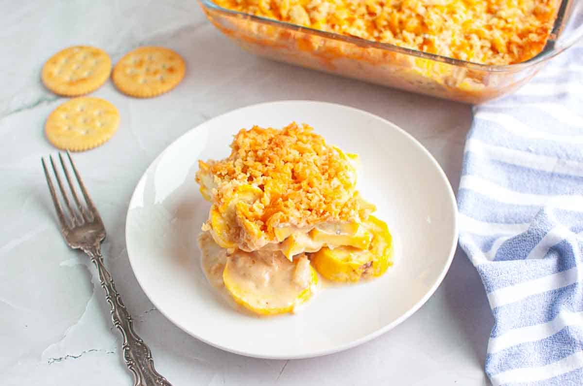 A serving of yellow squash casserole with a crispy crumb topping on a white plate, next to a fork, crackers, and a glass baking dish.