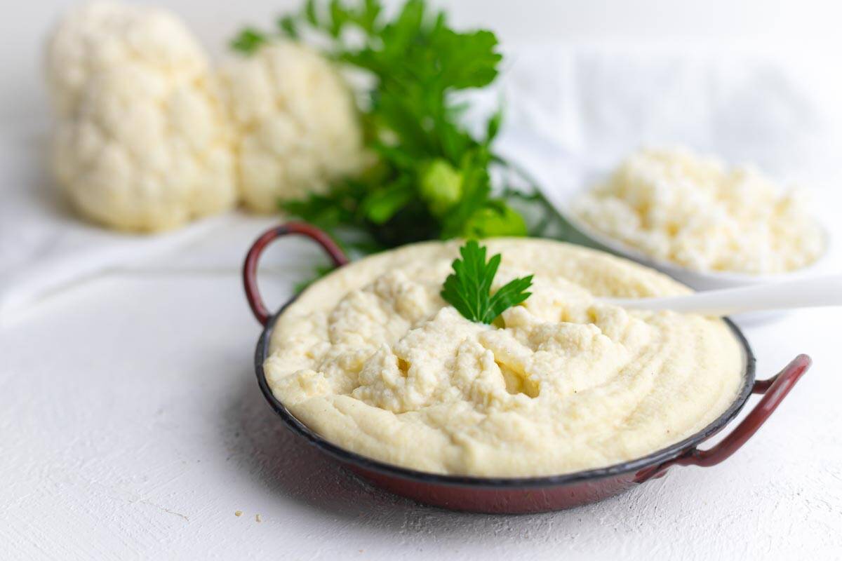 A bowl of creamy mashed cauliflower garnished with parsley, with raw cauliflower, parsley, and riced cauliflower in the background.