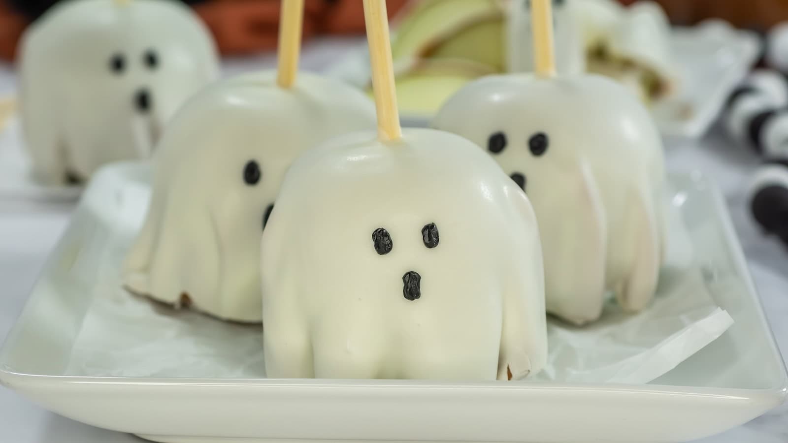 Three white-covered caramel apples on a square white plate, each decorated with eyes and mouth and skewered with a wooden stick in the middle. Behind the plate is another white apple on parchment paper.