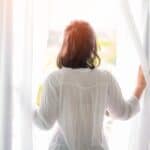 A person with dark hair, wearing a white shirt, stands facing a bright window, holding open white curtains to welcome the morning light during daylight saving time.