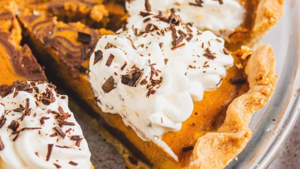 A close-up of pumpkin pie slices topped with whipped cream and chocolate shavings in a pie dish.