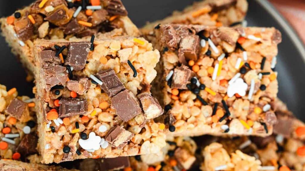 Close-up of rice crispy treats topped with chopped chocolate, orange and black sprinkles, and small white candy shapes, arranged on a dark plate.