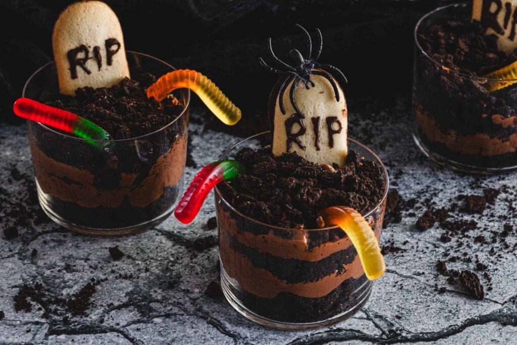 Three cups of layered chocolate pudding and crushed cookies decorated with candy worms, a cookie gravestone labeled "RIP," and a plastic spider on a gray stone-like surface.