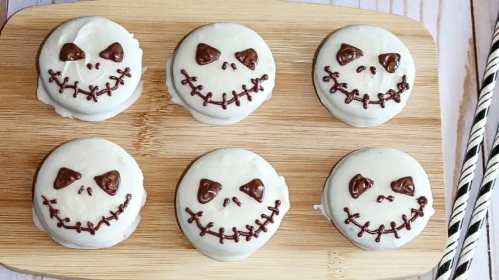 Six round cookies coated in white icing with chocolate decorations resembling stitched Jack Skellington faces, arranged on a wooden board.
