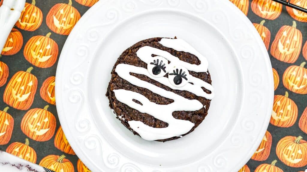 A round chocolate cookie with white icing stripes and two candy eyes on a white plate, set on a pumpkin-themed tablecloth.