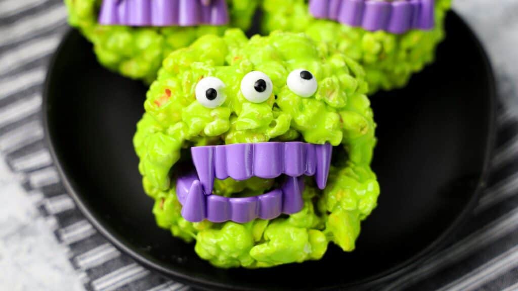 Green monster-shaped treat with candy eyes and purple plastic fangs, placed on a black plate on a striped cloth background.