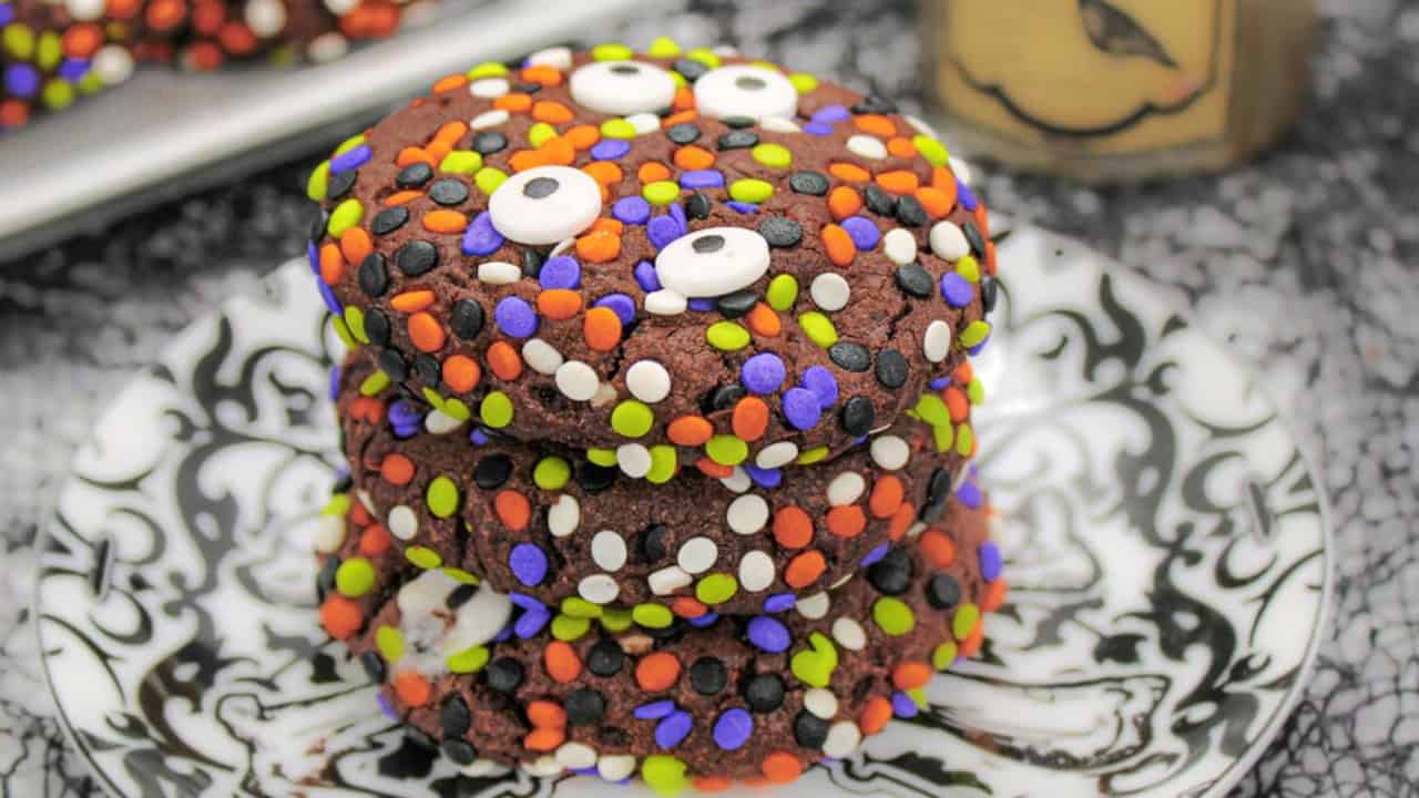 A stack of three chocolate cookies covered in colorful sprinkles, including orange, purple, green, white, and black, with candy eyes. They are on a decorative black and white plate with a patterned cloth in the background.