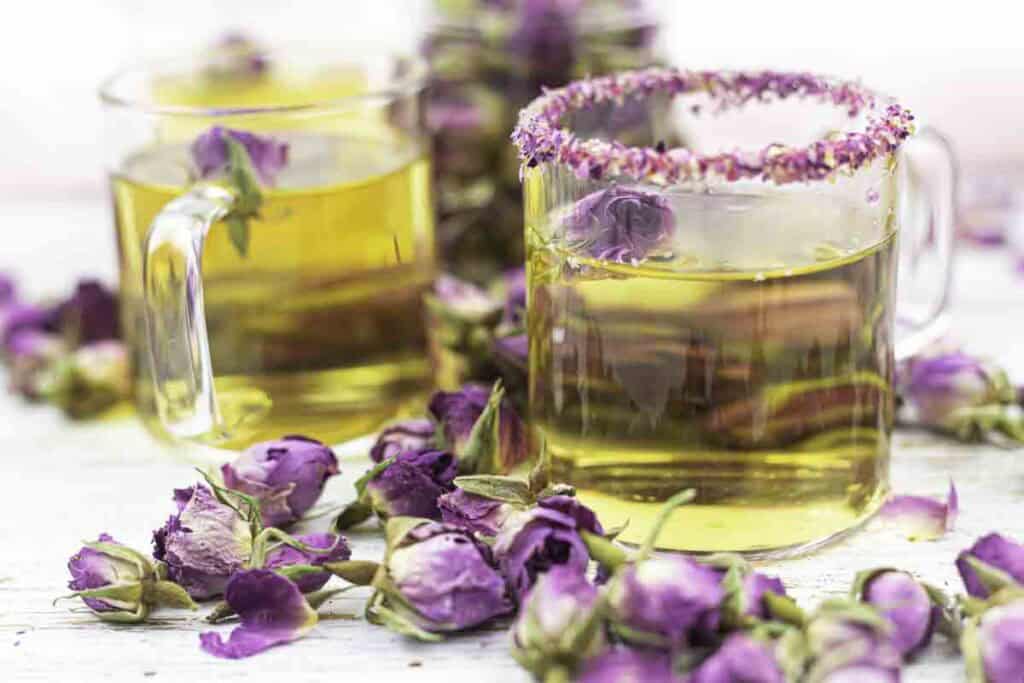 Two glass mugs of herbal tea with dried rosebuds, one mug has a rose petal rim; loose rosebuds are scattered on a white surface around the cups.