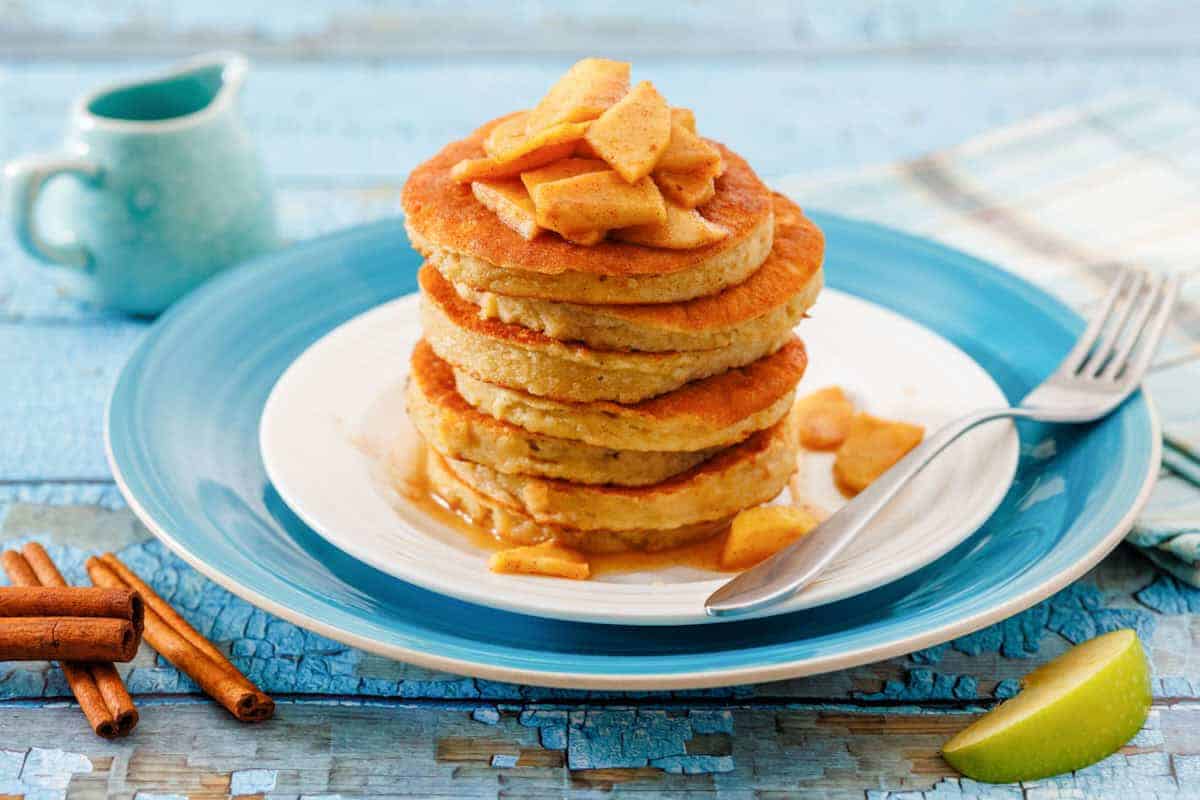 A stack of pancakes topped with cooked apple slices and syrup on a white plate with a blue rim, next to a fork, cinnamon sticks, and a small jug.