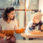 A young woman sits beside an older woman eating at a table, smiling and touching her arm in a friendly gesture in a bright indoor setting.
