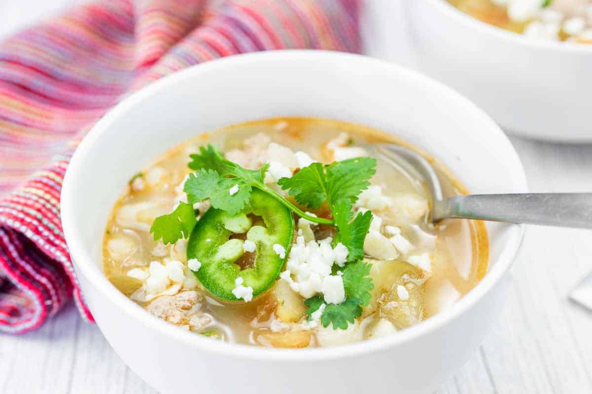 A bowl of Green pork posole soup garnished with cilantro, sliced jalapeño, and crumbled cheese, with a spoon resting inside and a striped cloth in the background.
