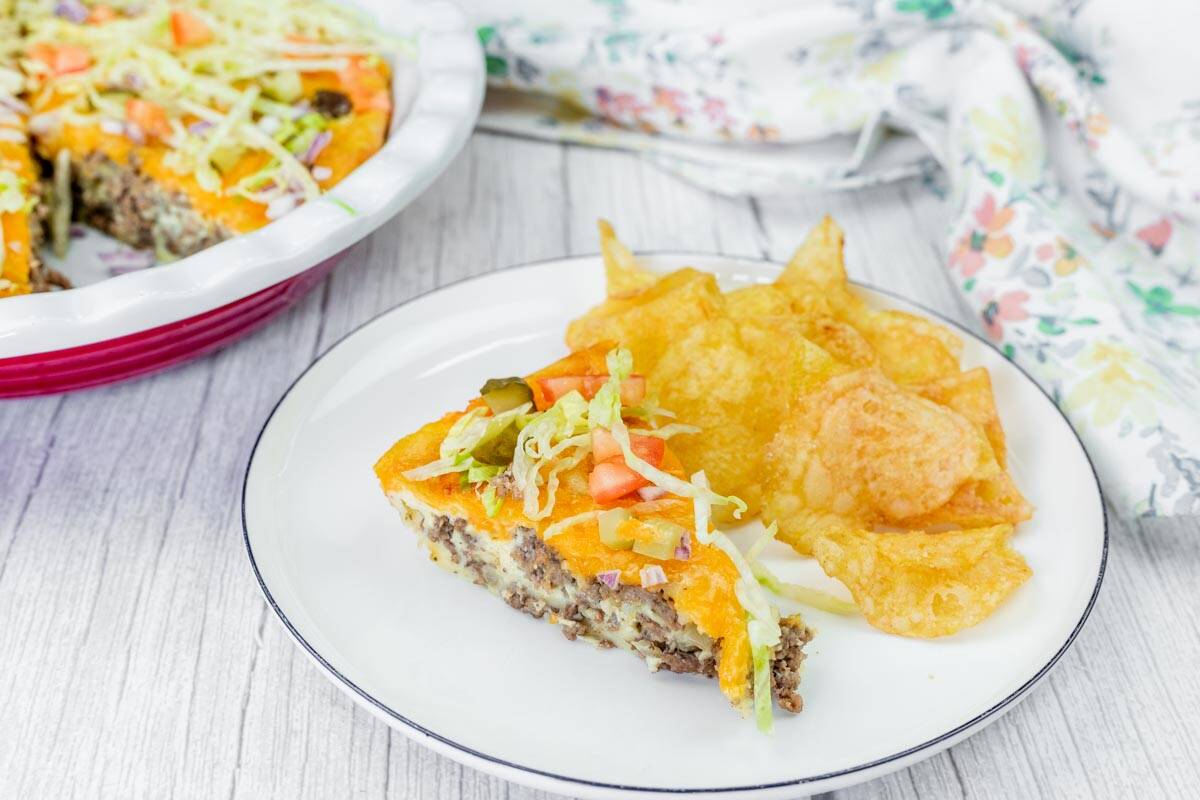 A plate with a slice of Beefy Cheeseburger Pie topped with shredded lettuce and tomatoes, served with potato chips.