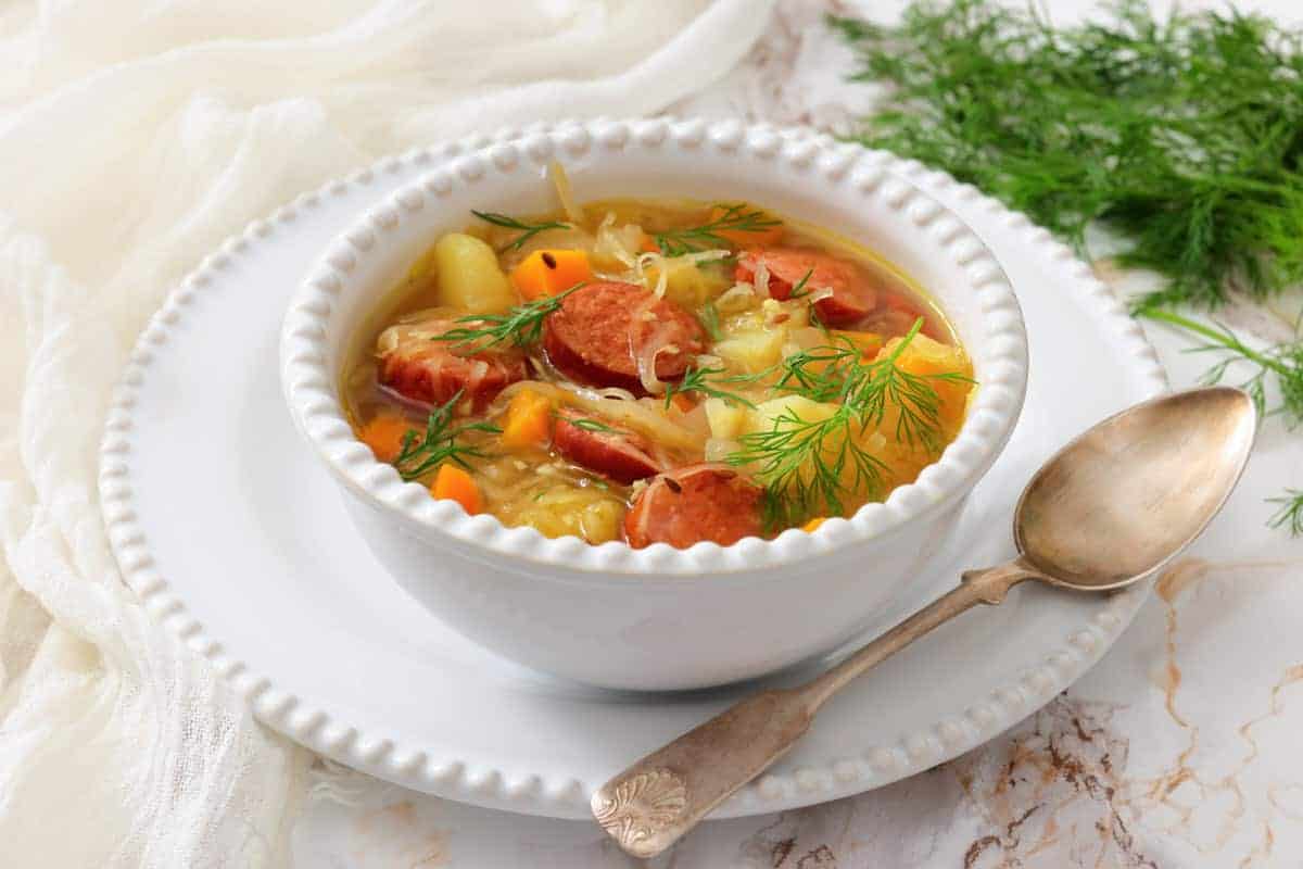 A white bowl of soup with sausage, potatoes, carrots, and dill garnish sits on a plate with a spoon on a marble surface.