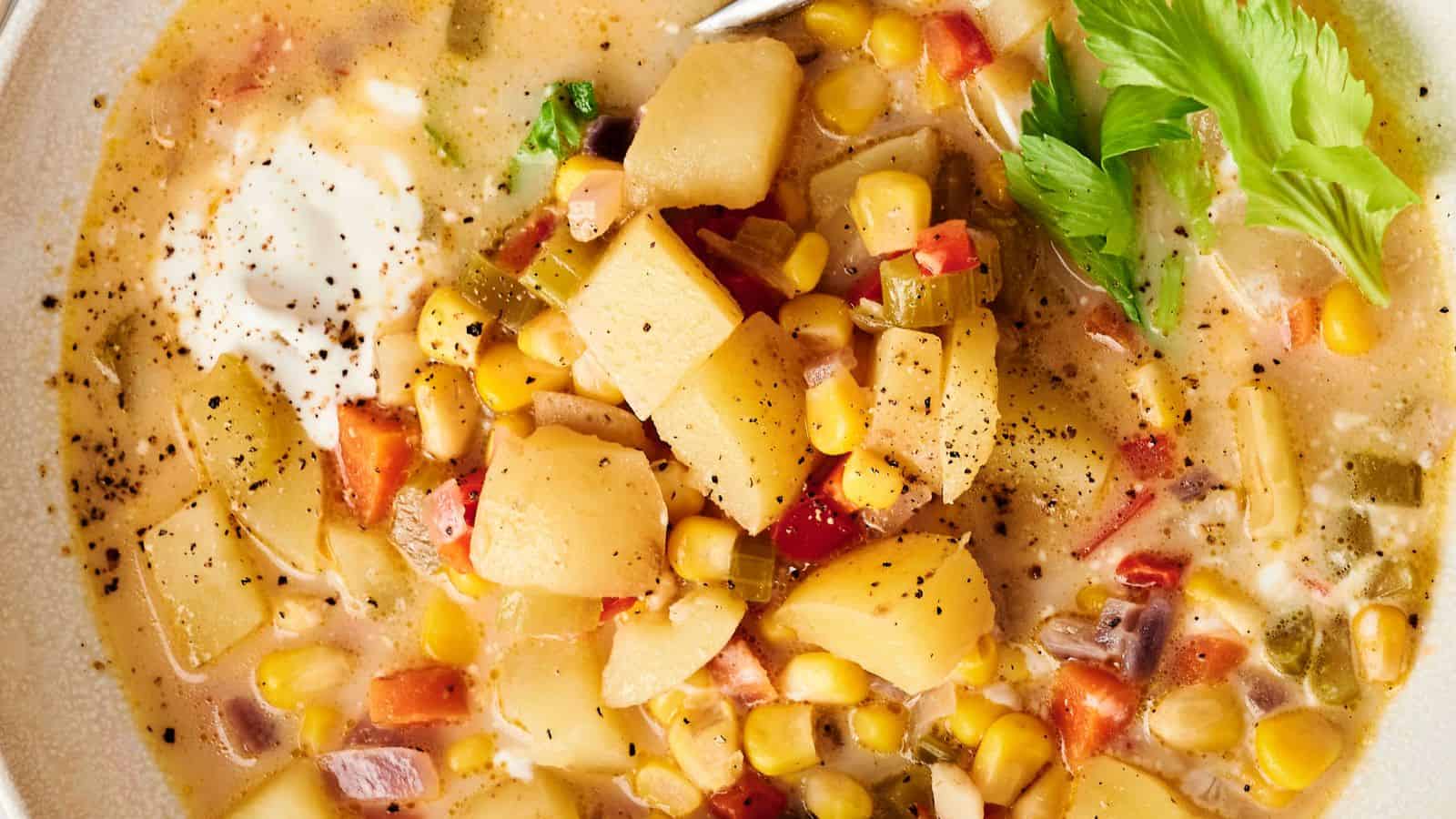 A bowl of vegetable chowder with potatoes, corn, celery, and peppers, topped with black pepper and a dollop of sour cream, garnished with a celery leaf.