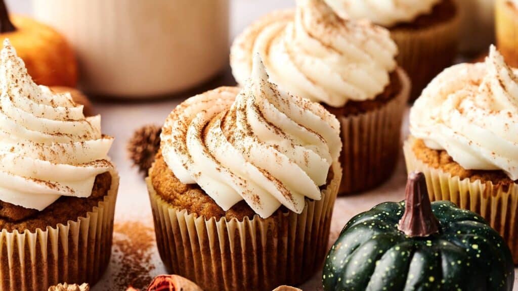 Pumpkin cupcakes with swirled cream cheese frosting and a sprinkle of cinnamon are arranged on a table next to a small decorative gourd.