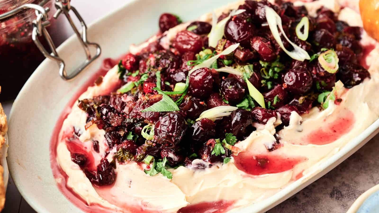 A platter of whipped cheese topped with roasted cranberries, chopped herbs, and sliced green onions.