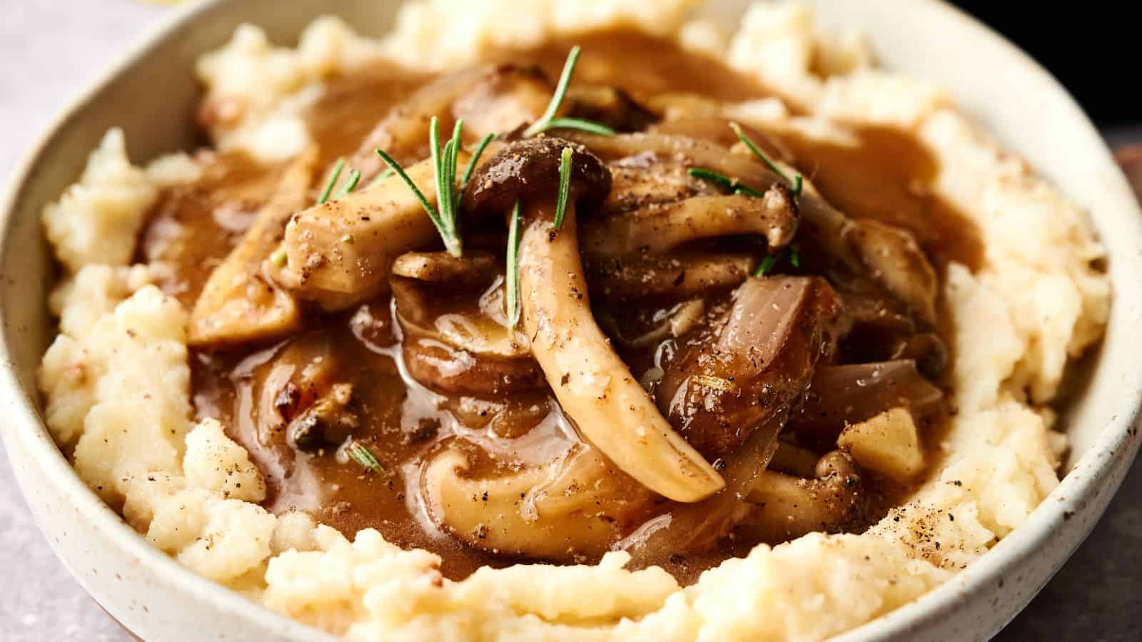 A bowl of mashed potatoes topped with mushroom and onion gravy, garnished with rosemary.