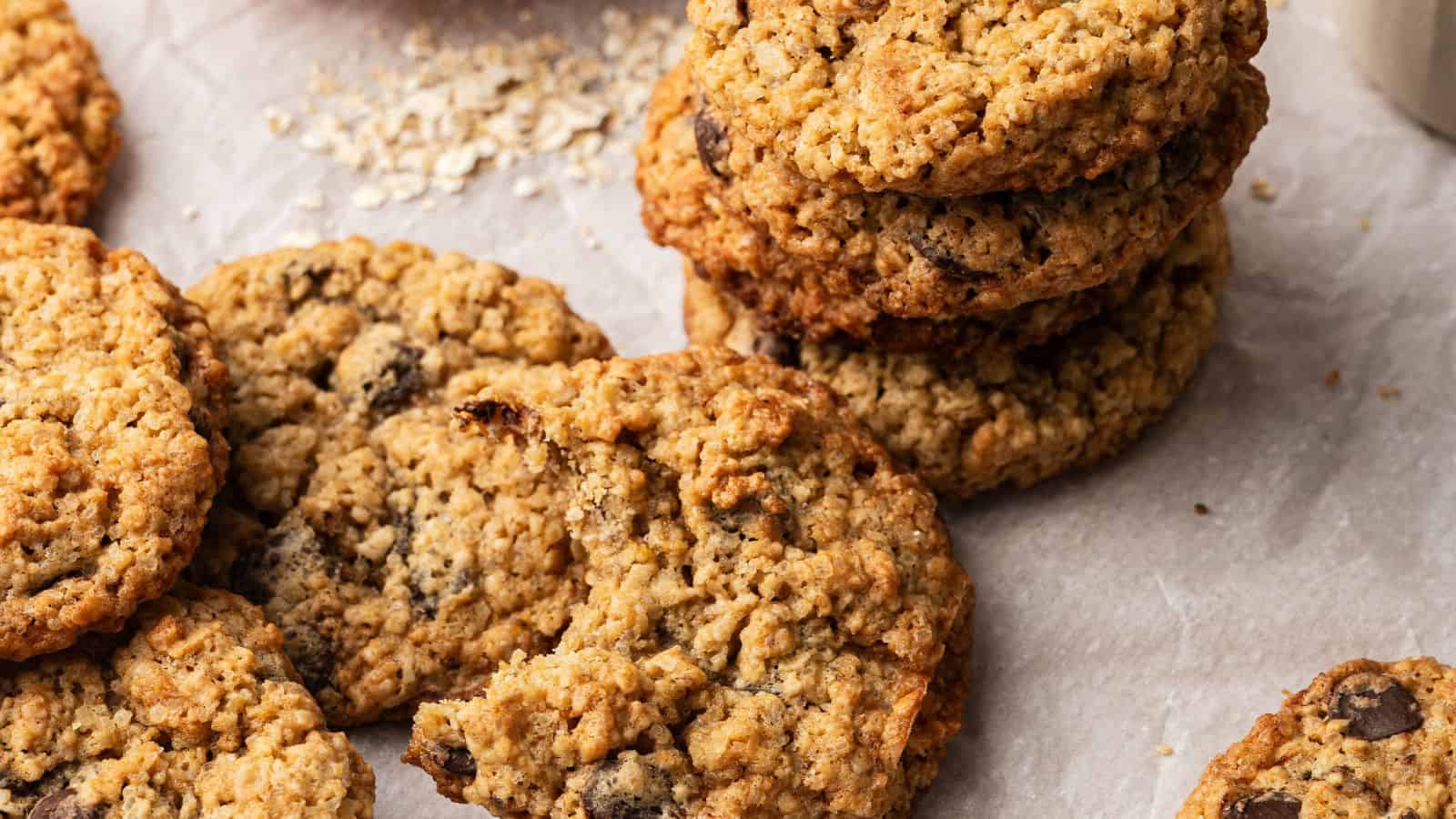 A stack and several scattered oatmeal chocolate chip cookies are shown on parchment paper, with oats visible in the background.