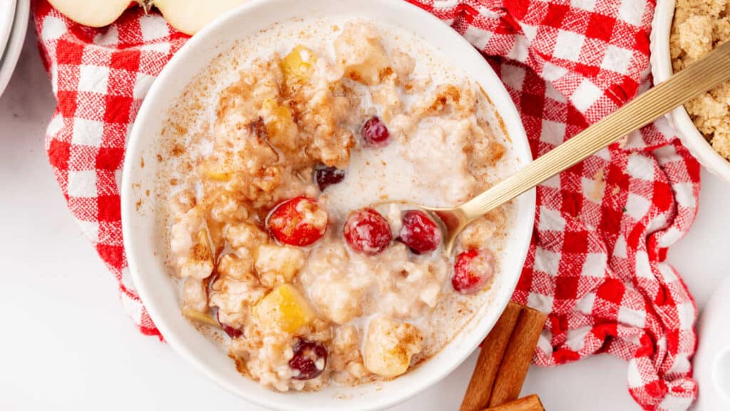 A bowl of oatmeal with cranberries and apples, placed on a red and white checkered cloth with a gold spoon and cinnamon sticks nearby.