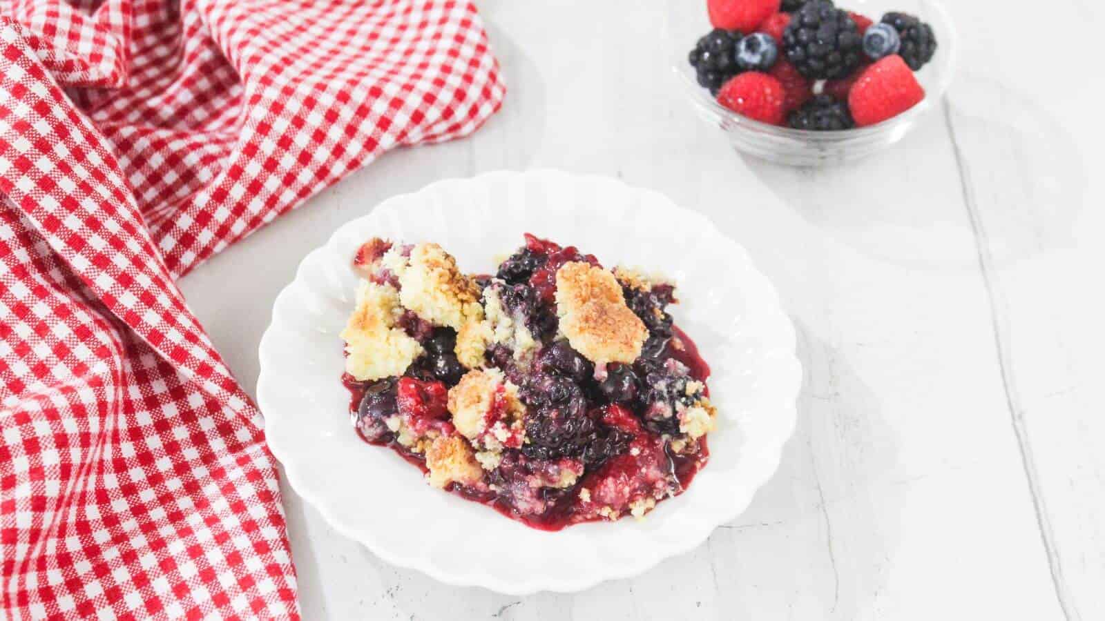 A serving of berry crumble on a white plate next to a red checkered cloth, with a small bowl of mixed berries in the background.
