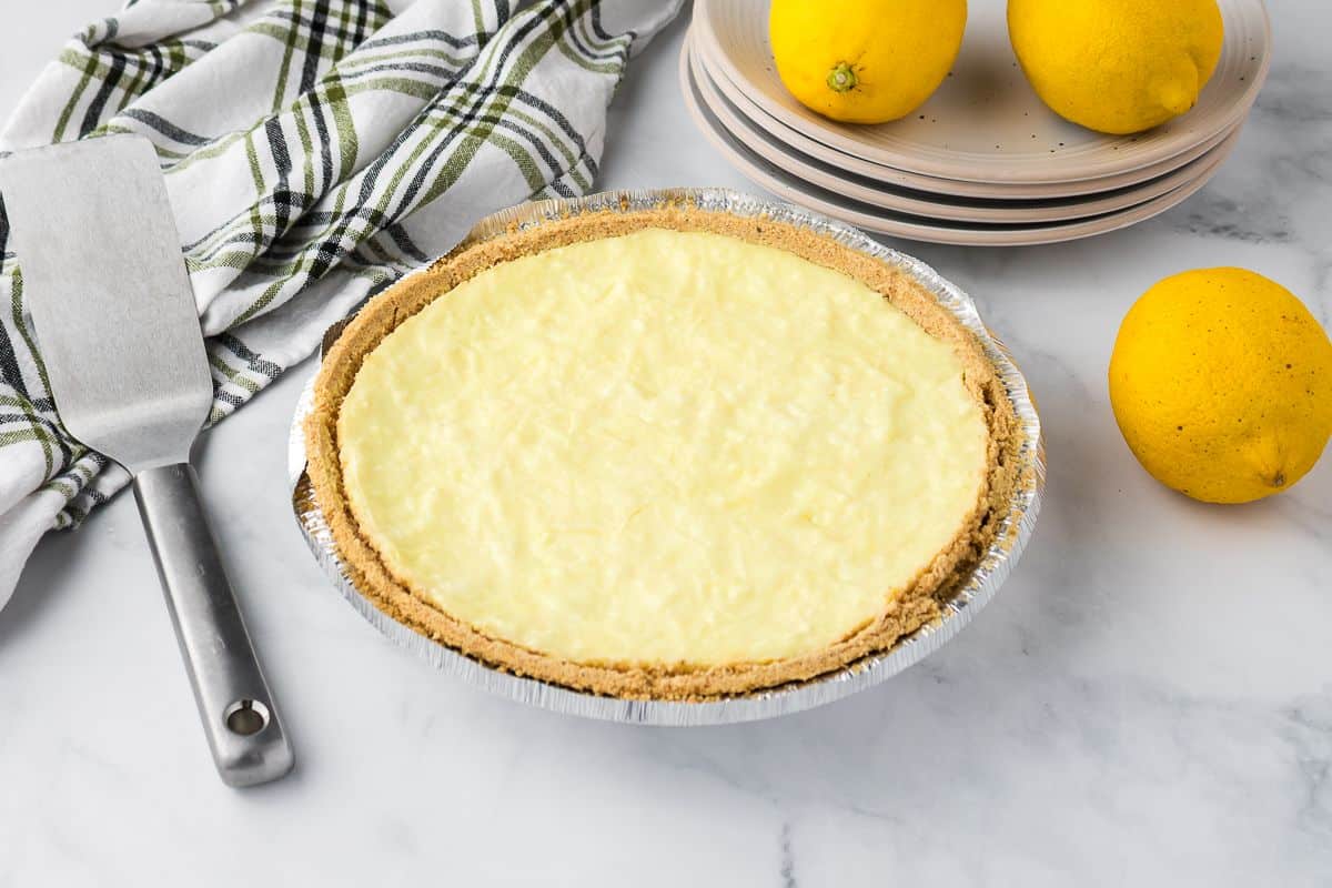 A lemon pie with a graham cracker crust sits on a marble surface next to a spatula, a striped kitchen towel, stacked plates, and three whole lemons.