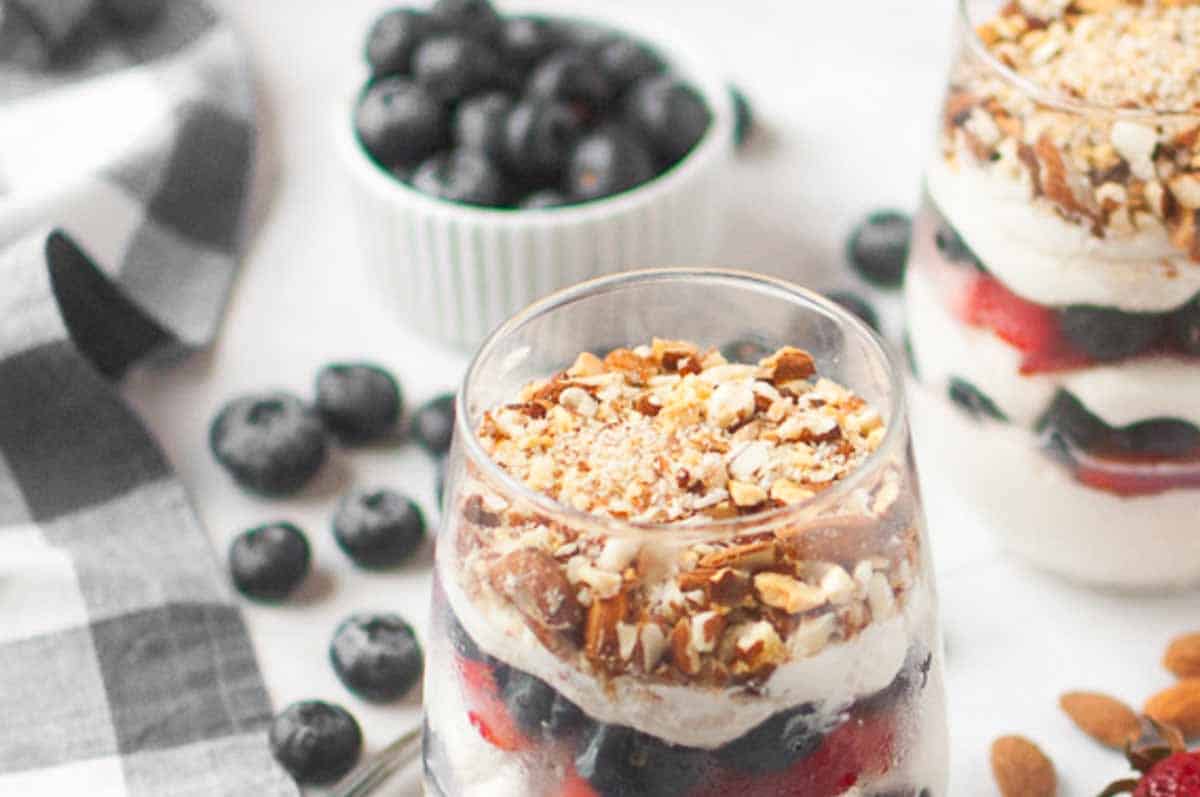 A glass filled with layers of cheesecake and berries sits on a table, surrounded by scattered blueberries and a bowl of blueberries in the background.