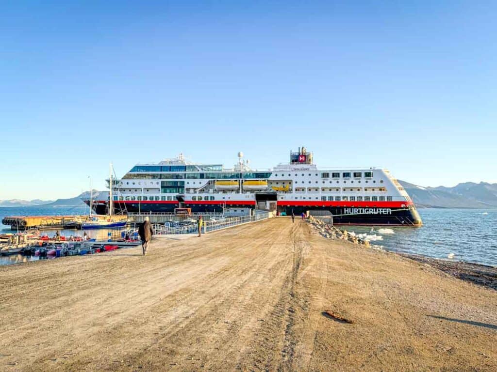 A large Hurtigruten cruise ship is docked at a pier beside smaller boats, with a person strolling along the pier and the dramatic mountains of Norway&rsquo;s fjords rising in the background.