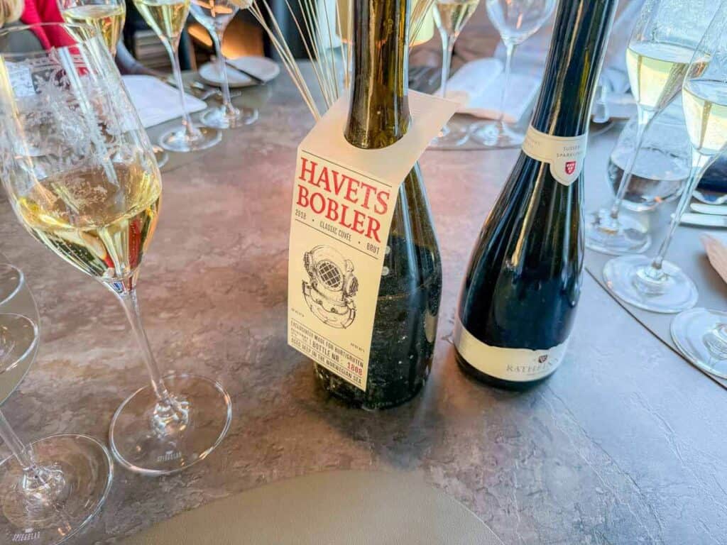 Two bottles of sparkling wine are on a table surrounded by glasses filled with sparkling wine, evoking the shimmering elegance of Norway&rsquo;s fjords. One bottle has a label that reads "HAVETS BOBLER.