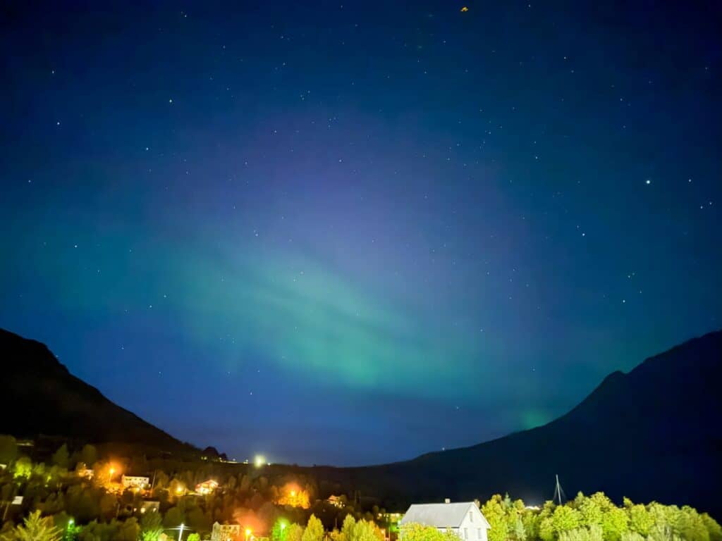 Northern lights glow faintly in the night sky above a small town nestled among illuminated houses, surrounding mountains, and the serene beauty of Norway&rsquo;s fjords.