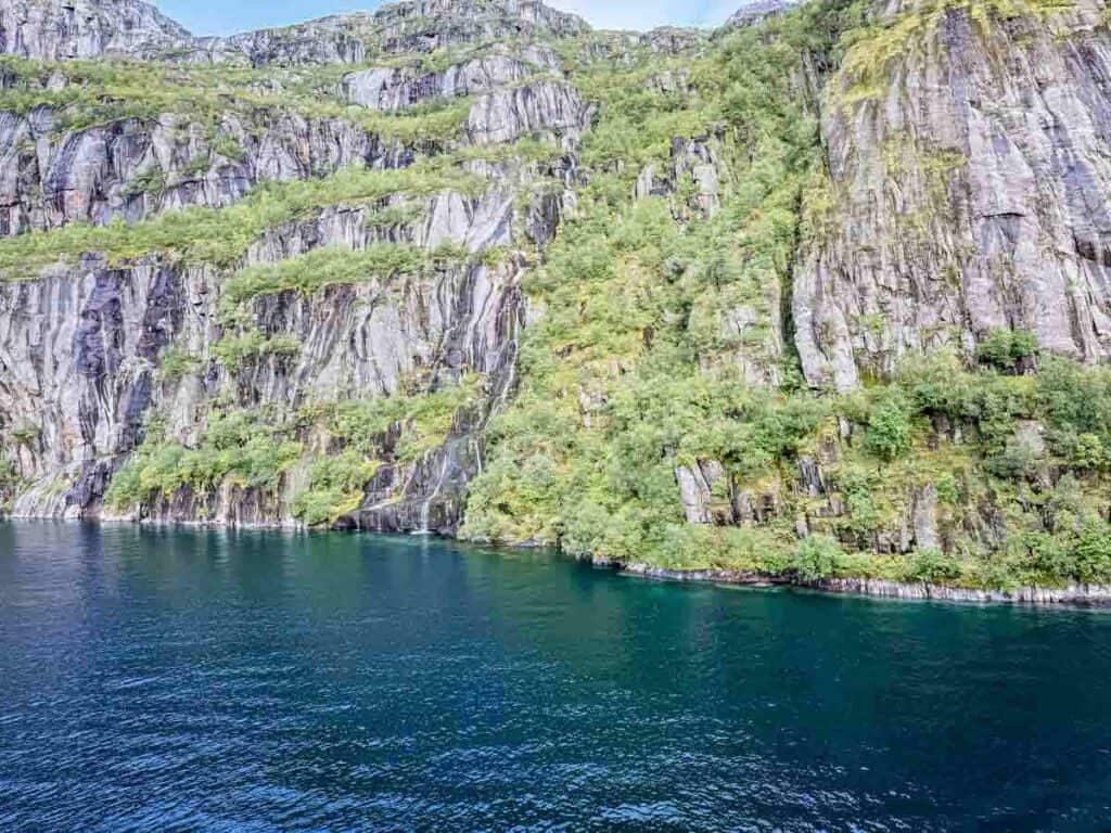 Steep rocky cliffs covered with greenery rise from the edge of calm blue water under a partly cloudy sky, capturing the breathtaking beauty of Norway&rsquo;s fjords.