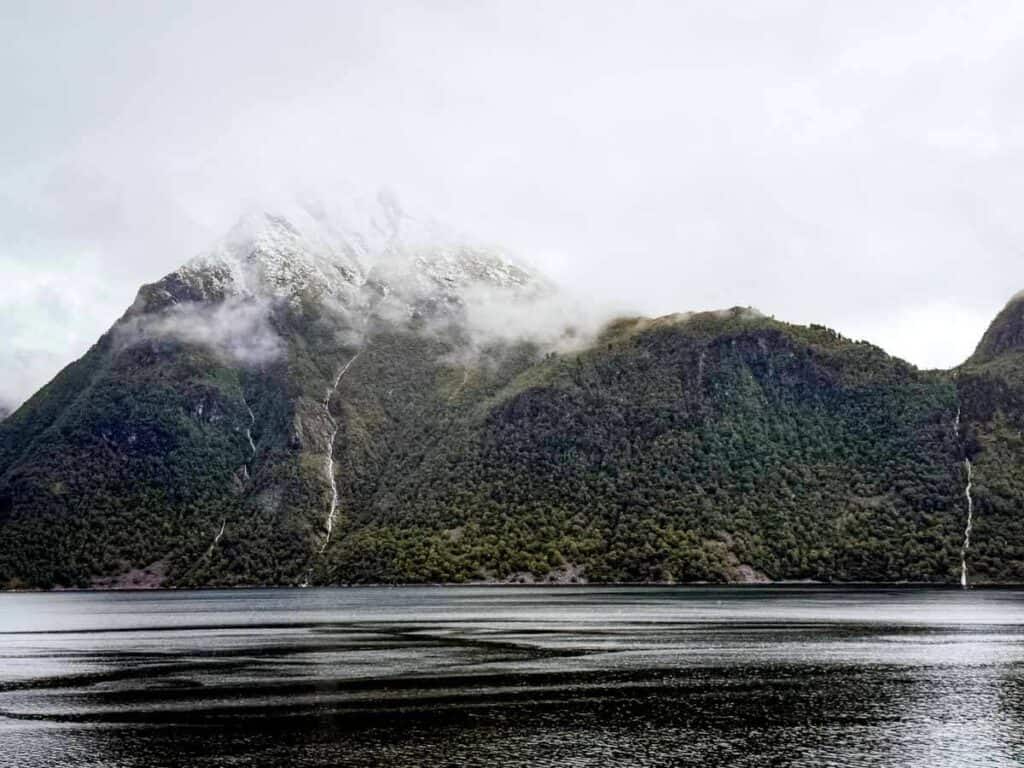 Snow-capped mountain partially covered by clouds, with green slopes and small waterfalls, reflected in a calm body of water&mdash;capturing the serene beauty typical of Norway&rsquo;s fjords.