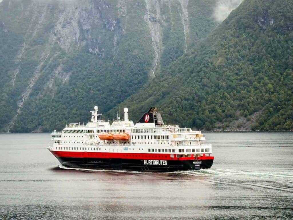 A Hurtigruten cruise ship sails through calm water near tree-covered mountains and Norway&rsquo;s fjords under a cloudy sky.