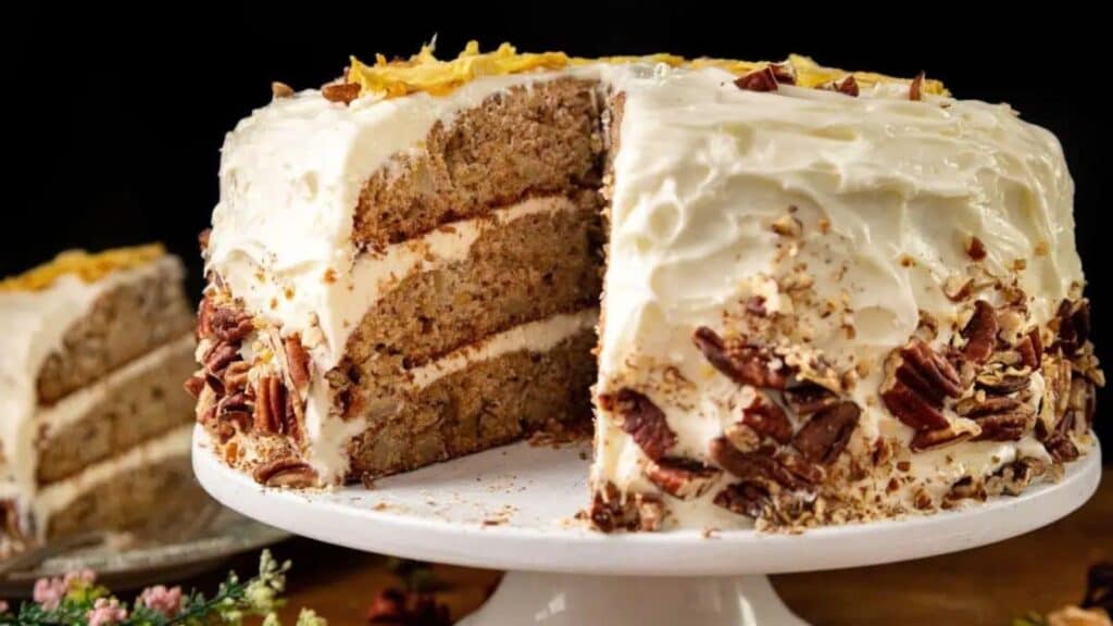 A three-layer cake with white frosting, topped and garnished with pecan halves and chopped pecans, sits on a white cake stand with a large slice removed.