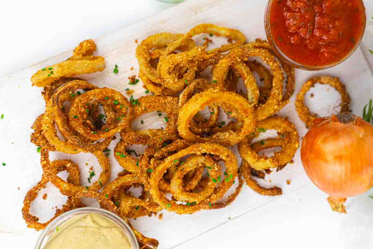 A pile of crispy onion rings garnished with herbs on a white surface, accompanied by dipping sauces and a whole onion.