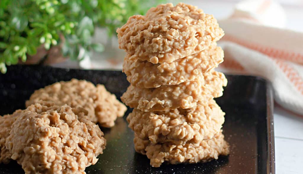 A stack of no-bake oatmeal cookies on a black tray, with a few separate cookies and a green plant in the background.