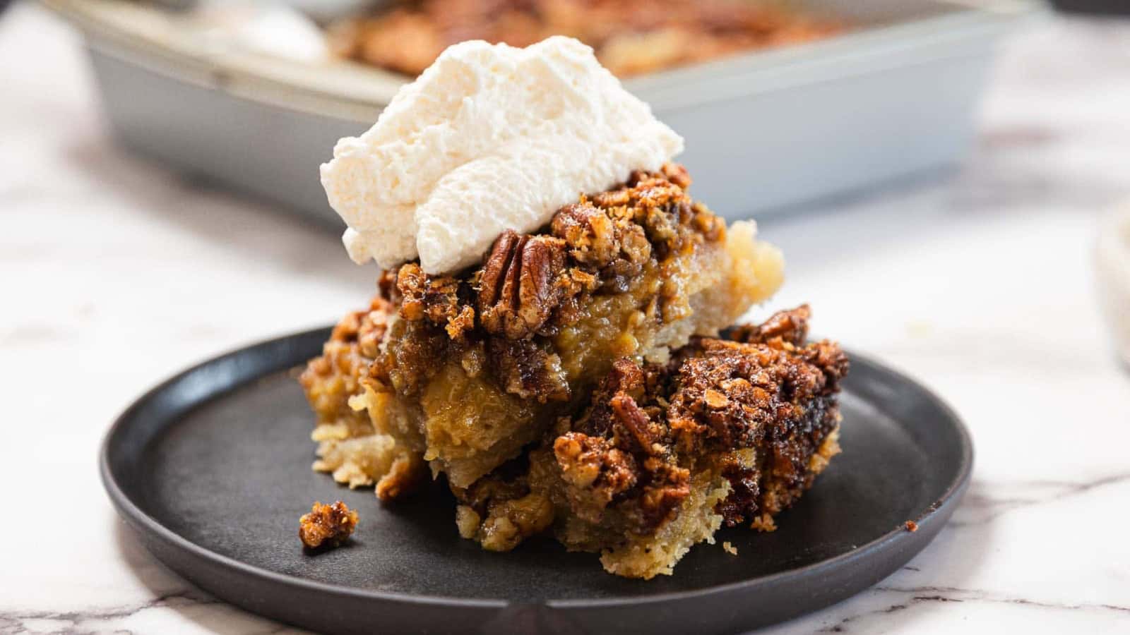 A slice of pecan pie topped with whipped cream on a black plate, with a baking dish in the background.