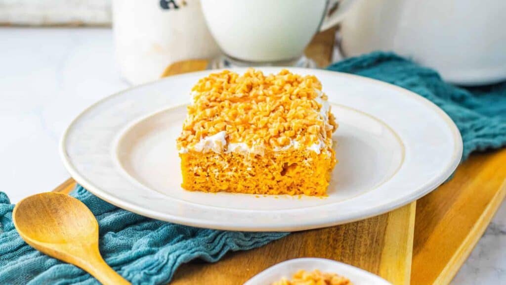 A square piece of cake with white frosting and crushed nuts on top, served on a white plate with a wooden spoon and a glass of milk in the background.