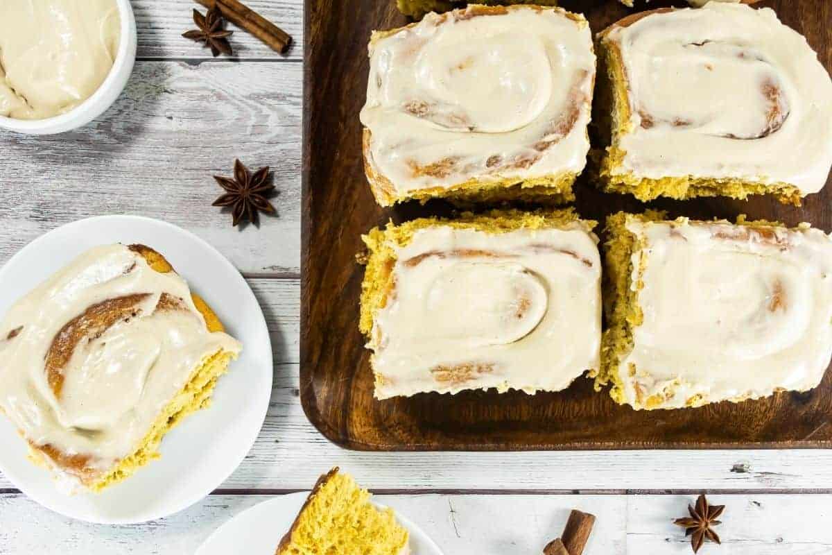 Four pumpkin cinnamon rolls are on a wooden board, with one roll served on a white plate.
