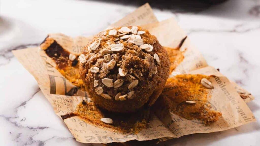 A muffin topped with oats sits on a piece of parchment paper with printed text, placed on a marble surface.