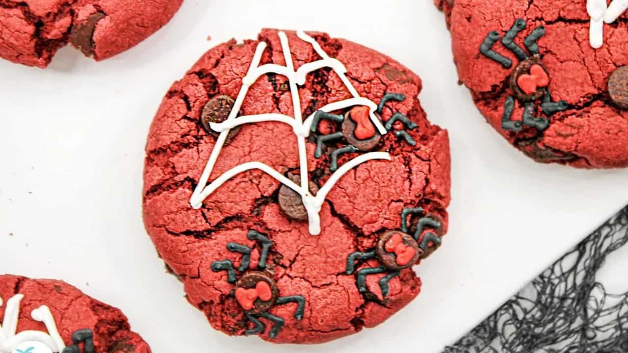 A red cookie decorated with white icing in a spiderweb pattern and black icing spiders with red details. Other similarly decorated cookies are partially visible around it.