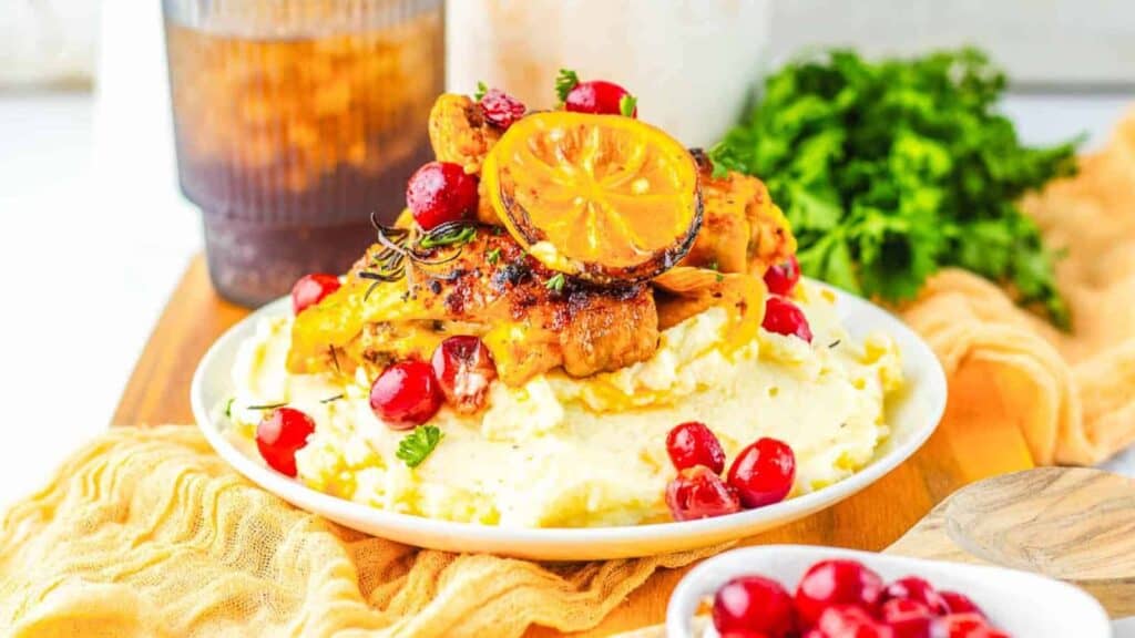 A plate of mashed potatoes topped with roasted chicken, a grilled lemon slice, red berries, and herbs, with a drink and fresh parsley in the background.